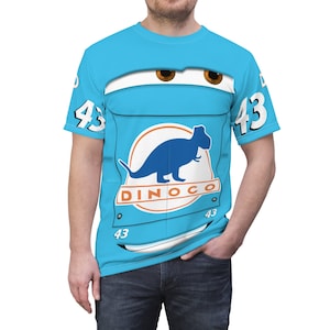 Può includere: Una maglietta blu con un design di auto da cartone animato. L'auto ha un logo di dinosauro che dice "Dinoco" e il numero "43" sul lato.