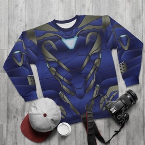 May include: A blue sweatshirt with a gold and silver metallic print of a superhero's chest armor. The sweatshirt is laid flat on a woodgrain surface with a camera, a hat, and a cup.