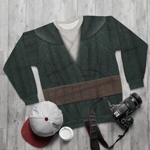 Op de afbeelding: Een groene sweatshirt met een bruin riemontwerp, een grijze en bordeauxrode baseballcap, een camera en een witte koffiekop. De sweatshirt heeft een gewatteerd patroon en een gelaagde kraag. De items zijn gerangschikt op een houten oppervlak.