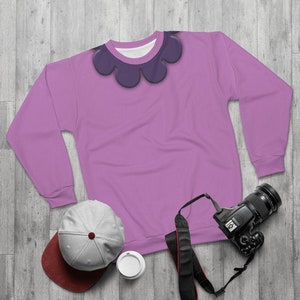 May include: A lavender sweatshirt with a purple flower design around the neckline. The sweatshirt is laying flat on a woodgrain surface with a gray baseball cap, a camera, and a white cup.