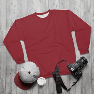 Op de afbeelding: Een rood rondhals sweatshirt met lange mouwen, een grijze baseballpet met een rode klep, een zwarte camera met een band en een witte beker op een grijze houten achtergrond.