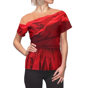 Könnte beinhalten: Rotes T-Shirt mit einem Aufdruck, der ein rotes Kleid mit drapiertem, schulterfreiem Ausschnitt und einem Gürtel in der Taille zeigt. Das Shirt hat kurze Ärmel und einen beigefarbenen Mesh-Einsatz am Ausschnitt und an den Schultern. Das Model trägt eine schwarze Hose.