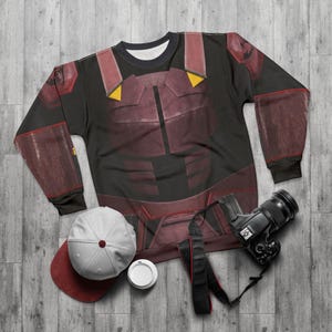 Peut inclure: Sweat-shirt noir avec un motif bordeaux et jaune ressemblant à une armure. Une casquette de baseball blanche et bordeaux, un appareil photo et une tasse à café sont à côté du sweat-shirt sur une surface en bois.