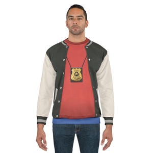 May include: A person wearing a red shirt with a gold police badge and a black and white letterman jacket. The badge is on a chain and has the words "Sergeant's Police" on it. The jacket has white sleeves and a black body.