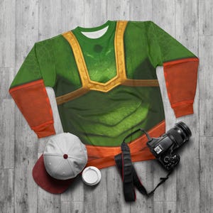 May include: Green and orange sweatshirt with a detailed design. The sweatshirt features a green torso with gold-colored accents and orange sleeves. A white baseball cap, a camera, and a coffee cup are also visible.