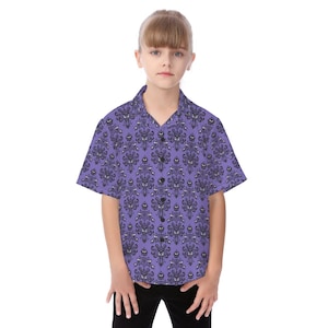 Pode incluir: Uma camisa de manga curta com fundo roxo e um padrão repetido de desenhos de casas assombradas pretas e estilizadas. A camisa, usada por uma pessoa, tem uma gola clássica e fecho de botões.
