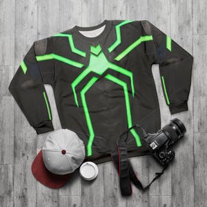 Op de afbeelding: Zwart sweatshirt met een neon groen spinnenweb ontwerp. Een grijze en bordeauxrode baseball cap, een witte koffiebeker en een zwarte camera met een riem staan ook op de foto. De items liggen op een grijze houten ondergrond.