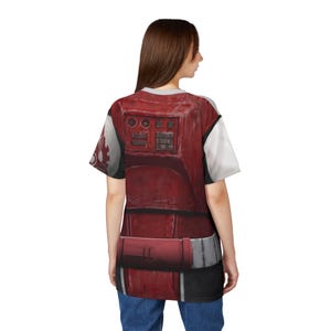 May include: A red and white t-shirt with a detailed armour design. The design includes rectangular panels, buttons, and a belt, with a weathered appearance. The sleeves are white. The shirt is worn by a person.