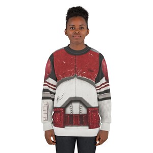 May include: A crew neck sweatshirt with a design in red, white, and grey, resembling a military uniform. The long-sleeved top has a detailed graphic print across the chest and shoulders.