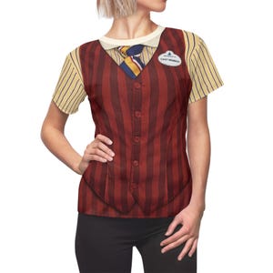Könnte beinhalten: Ein kurzärmeliges T-Shirt mit einem Aufdruck eines rot-braun gestreiften Westen mit Knöpfen, einem weißen Kragenhemd und einer gestreiften Krawatte. Das T-Shirt hat auch ein Namensschild mit der Aufschrift "CAST MEMBER". Die Ärmel sind gelb und weiß gestreift.