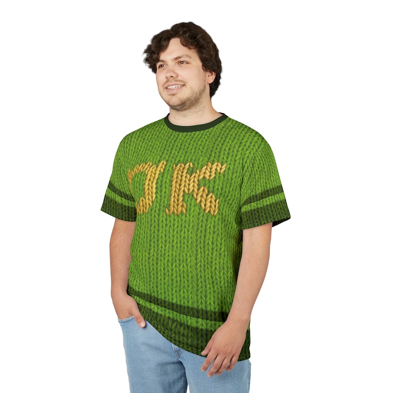 K&ouml;nnte beinhalten: Gr&uuml;nes Kurzarm-T-Shirt mit Strickmuster und den Buchstaben "JK" in Gold. Das Shirt hat schwarze Streifen an den &Auml;rmeln und am Kragen. Die Person tr&auml;gt eine blaue Jeans.