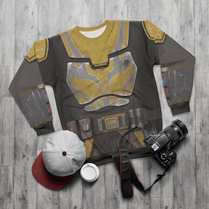 May include: A black and grey sweatshirt featuring a yellow and silver armour design. The long-sleeved shirt has a belt design with pouches. A white and maroon baseball cap, a white coffee cup, and a black camera are also in the image.