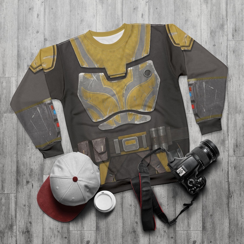 May include: A black and grey sweatshirt featuring a yellow and silver armour design. The long-sleeved shirt has a belt design with pouches. A white and maroon baseball cap, a white coffee cup, and a black camera are also in the image.