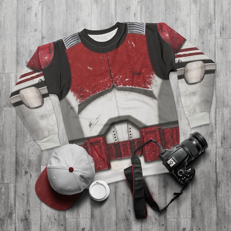May include: A long-sleeved shirt with a red and white armour design, a white and red baseball cap, a black camera with a strap, and a white coffee cup. The shirt has a distressed appearance.