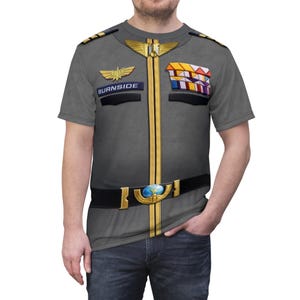 Può includere: T-shirt grigia con un design stampato che ricorda una divisa militare. Il design include dettagli dorati, una cintura nera con fibbia a globo e il nome "BURNSIDE" su una toppa. Maniche corte e girocollo.