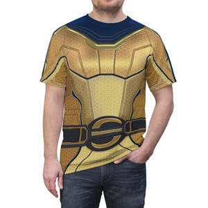 May include: Gold and navy blue t-shirt featuring a detailed design resembling a superhero costume. The shirt has a textured pattern, a black belt with a gold buckle, and a navy blue neckline and shoulder accents. The short-sleeved shirt is made of a soft material.