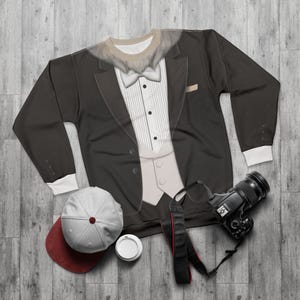May include: A black sweatshirt with a tuxedo design, featuring a white dress shirt, bow tie, and vest. A white and maroon baseball cap, a camera, and a coffee cup are also visible on a wooden surface.