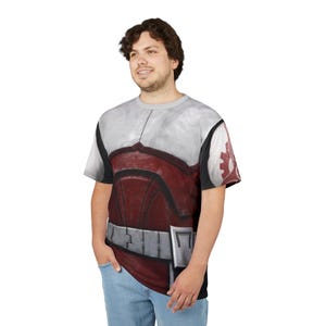 May include: A short-sleeved t-shirt with a detailed graphic design. The shirt displays a red and white armour-like pattern across the chest and shoulders, with a silver belt design at the waist. The design includes a circular emblem on the sleeve.