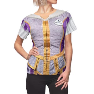 Könnte beinhalten: Ein kurzärmeliges T-Shirt mit einem lila-goldenen Miederdesign, das an ein Prinzessinnenkostüm erinnert. Das Shirt hat einen weißen Kragen und Ärmel mit einem "CAST MEMBER" Namensschild. Das Design beinhaltet florale Muster und dekorative Verzierungen.