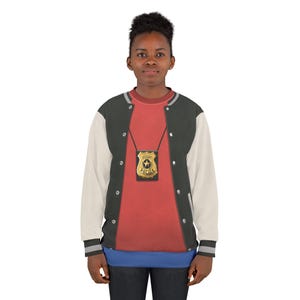 May include: A person wearing a black, white, and gray letterman jacket over a red shirt. A gold-colored police badge hangs from a black lanyard. The jacket has white sleeves and a gray and black striped hem.