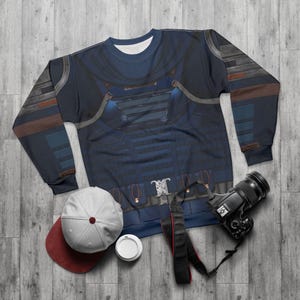May include: A navy blue sweatshirt with a detailed graphic design resembling armor. The design incorporates shades of blue, gray, and brown. A white and maroon baseball cap, a disposable coffee cup, and a black camera with a strap are also visible.