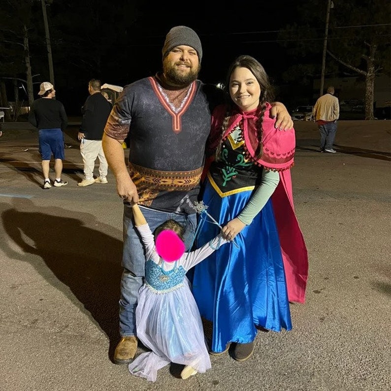May include: A family dressed in costumes for a Halloween event. A man wearing a dark patterned shirt with a beard is holding a young girl dressed as Elsa from Frozen. A woman wearing a pink and blue dress with a long cape is holding the girl's hand. The girl is wearing a blue and white dress with a white cape.