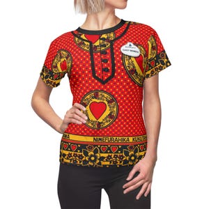 Könnte beinhalten: Rotes T-Shirt mit einem sich wiederholenden Muster aus gelben Kreisen, Herzen und schwarzen floralen Designs. Das Shirt hat einen schwarzen Kragen und Besatz, mit einem "CAST MEMBER" Abzeichen. Der Text "NIMEFURAHIKA KUKUTANA" ist unten aufgedruckt.