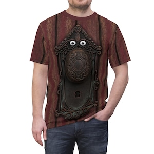 May include: A short-sleeved t-shirt featuring a graphic print of a dark red wooden door with a central ornate metal door handle and backplate. The door handle is designed with two white circular eyes positioned above the central knob.