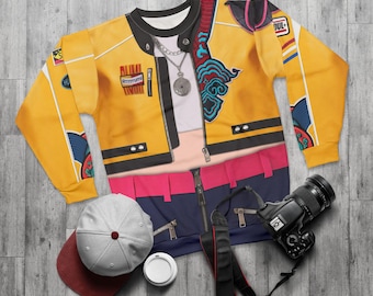 May include: A yellow long-sleeved shirt with a printed design resembling a jacket, featuring a zipper, pockets, and decorative patches. A gray baseball cap, a camera, and a coffee cup are also present.