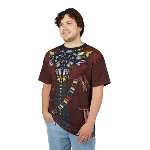 Puede incluir: Una camiseta marr&oacute;n con un dise&ntilde;o estampado de una chaqueta formal y una gran pajarita. El dise&ntilde;o de la chaqueta incluye un chaleco, una faja colorida y detalles florales. La camiseta la lleva una persona con vaqueros azules.