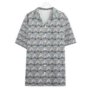 May include: A short-sleeved button-down shirt with a geometric pattern in shades of blue, tan, and gray. The shirt has a V-neck collar and a series of small white buttons down the front. The pattern is a repeating design of three-dimensional shapes.