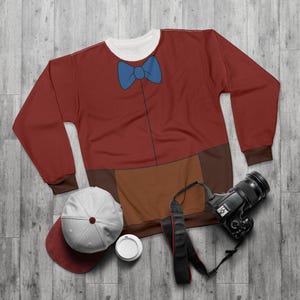 Könnte beinhalten: Ein weinrotes Sweatshirt mit einem blauen Fliegen-Design. Das langärmlige Shirt hat einen braunen unteren Teil und Manschetten mit einem weißen Rundhalsausschnitt. Eine Baseballkappe, eine Kaffeetasse und eine Kamera liegen neben dem Shirt.