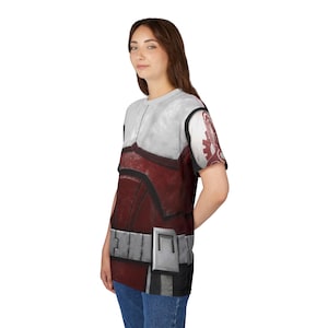 May include: A short-sleeved t-shirt with a print of a red and white armored suit. The design includes a silver belt and shoulder details, with a graphic on the right sleeve. The shirt is worn by a person.