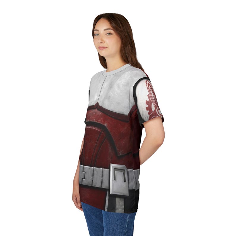 May include: A short-sleeved t-shirt with a print of a red and white armored suit. The design includes a silver belt and shoulder details, with a graphic on the right sleeve. The shirt is worn by a person.