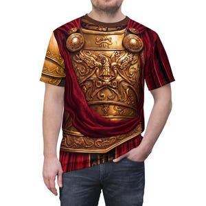 May include: A short-sleeved t-shirt featuring a detailed golden Roman armor design. The armor is adorned with intricate carvings and is draped with a flowing red fabric. The shirt has a brown neckline and is worn by a person.