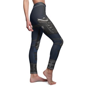 May include: Navy blue leggings with a detailed graphic print of a firearm and tactical gear. The leggings feature a high waistband and a form-fitting design, with accents of black and gray. The overall aesthetic suggests a military or tactical theme.