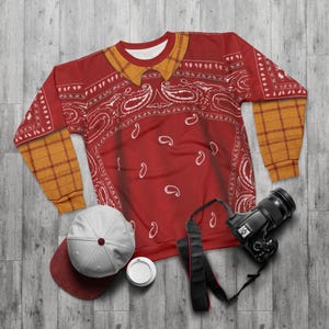 May include: A long-sleeve sweatshirt featuring a red bandana print with white paisley patterns, a yellow plaid collar, and yellow plaid sleeves. Beside the sweatshirt are a white baseball cap with a red brim and a black digital camera.