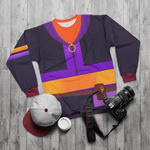 Könnte beinhalten: Ein schwarzer Pullover mit einem lila, orange und gelben Design. Das Design zeigt eine stilisierte Figur mit einem Gürtel und einer Tasche. Der Pullover hat lange Ärmel und einen Rundhalsausschnitt.