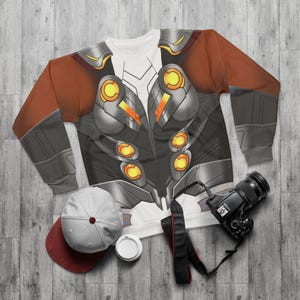 Puede incluir: Una sudadera de manga larga con un diseño gráfico detallado que se asemeja a una armadura. El diseño incorpora tonos de gris, naranja y amarillo. También se ven una gorra de béisbol gris y granate, una taza de café blanca y una cámara negra.