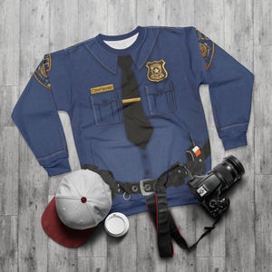 May include: A blue sweatshirt designed to look like a police uniform, complete with a badge, tie, and name tag that reads "Clawhauser." A baseball cap, coffee cup, and camera are next to the sweatshirt.