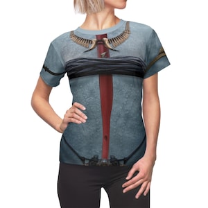 Pode incluir: T-shirt azul claro com um design gráfico detalhado. O design inclui uma faixa vertical vermelha, uma faixa horizontal preta e elementos decorativos na gola e nos ombros. A camisa tem mangas curtas e gola redonda.