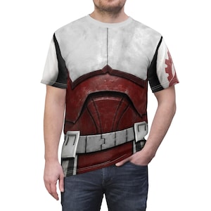 May include: A white t-shirt with a detailed graphic of red and white battle armour. The design includes a white chest plate, red armour, and a silver belt. The sleeves have black accents and a red and white emblem.