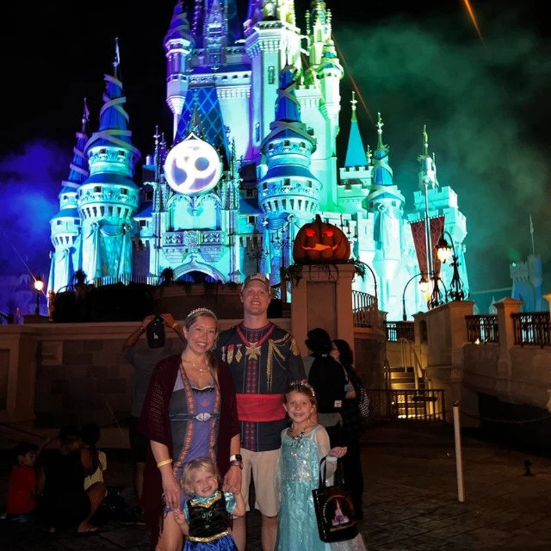 May include: A family of four dressed in Halloween costumes pose in front of Cinderella's Castle at Walt Disney World. The castle is lit up in blue and green lights, and there is a jack-o'-lantern in front of it. The family is dressed as characters from Disney films, including Elsa, Anna, and Aladdin.