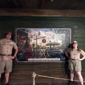 Pode incluir: Duas pessoas vestidas com roupas de safari cor c&aacute;qui est&atilde;o em frente a um grande cartaz que anuncia a Jungle Navigation Company Ltd. O cartaz apresenta uma ilustra&ccedil;&atilde;o colorida de uma cena de selva com um barco e um guindaste.