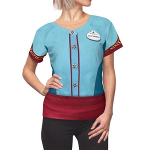 Könnte beinhalten: Ein hellblaues T-Shirt mit einem Mock-Button-Up-Design, mit einem roten Bund und Ärmelakzenten. Das Shirt enthält ein "CAST MEMBER" Namensschild. Das Shirt wird mit einer schwarzen Hose kombiniert.