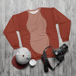 Könnte beinhalten: Ein rostfarbenes Sweatshirt mit hellbraunem Brustdesign, eine weiß-rote Baseballkappe, eine Kamera und eine Kaffeetasse. Das Sweatshirt hat einen Rundhalsausschnitt und lange Ärmel. Die Artikel sind auf einer Holzoberfläche angeordnet.