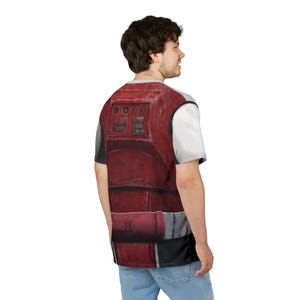 May include: A red and grey graphic t-shirt with a detailed armour design. The design includes rectangular panels, buttons, and a belt-like element. The shirt has a white short sleeve and a grey neckline.