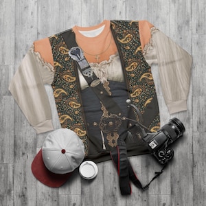 Op de afbeelding: Een crewneck sweatshirt met een piratenkostuumontwerp. Het ontwerp omvat een vest met een paisley patroon, een zwaard en een zilveren gesp. Een witte en rode baseballpet en een camera staan ook op de foto.