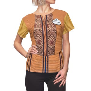 Könnte beinhalten: Ein kurzärmeliges, orange-gelbes T-Shirt mit einem dekorativen Muster auf der Vorderseite. Das Shirt hat ein vertikales Reißverschlussdesign und ein "CAST MEMBER" Namensschild. Die Ärmel sind einfarbig gelb mit kleinen Punkten.