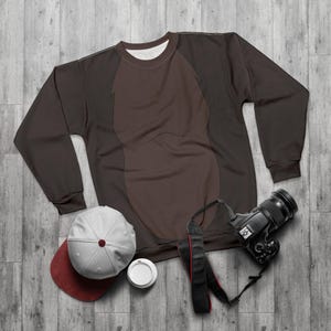 Könnte beinhalten: Ein braunes Sweatshirt mit einem hellbraunen Design auf der Vorderseite, eine grau-weinrote Baseballkappe, ein Einweg-Kaffeebecher und eine schwarze Kamera mit einem Riemen. Die Artikel sind auf einer grauen Holzoberfläche angeordnet.